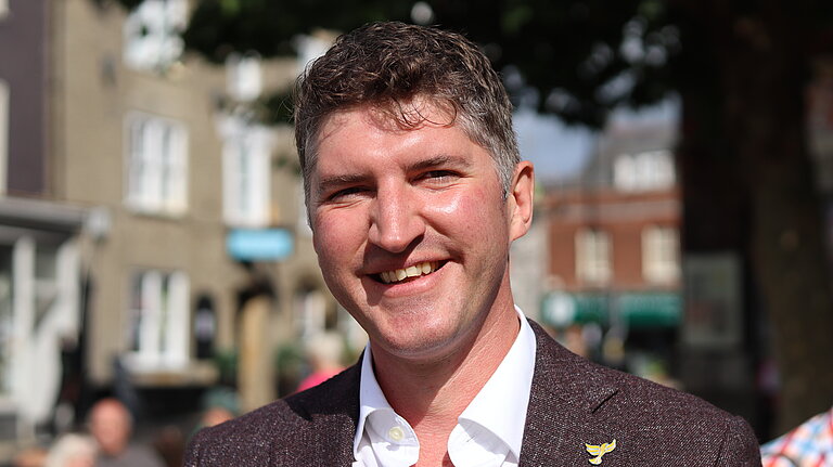 Edaward Morello wearing a white shirt and brown suit jacket, with a lib dem bird pin. he is stood in Buckydoo Square in Bridport