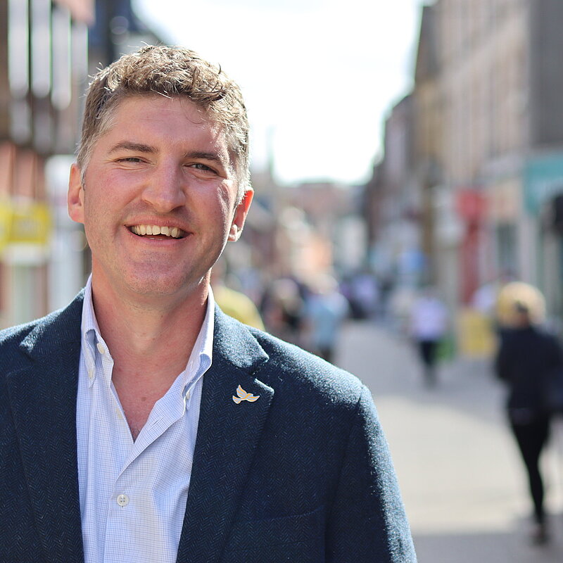 Edward Morello wearing a white shirt and navy suit jacket and a Lib Dem Bird Pin Badge, in Dorchester High Street