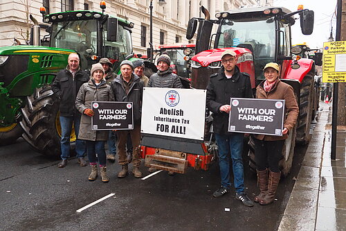 A group of farmers and supporters stand in front of large tractors on a city street, holding signs that read "WITH OUR FARMERS #together" and a banner stating "Abolish Inheritance Tax FOR ALL!" The individuals are dressed in outdoor work clothing, including jackets, hats, and boots.