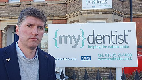Edward Morello's opinion of dentists in Dorset Lib Dem Candidate Edward Morello standing in front of an NHS dentist sign.