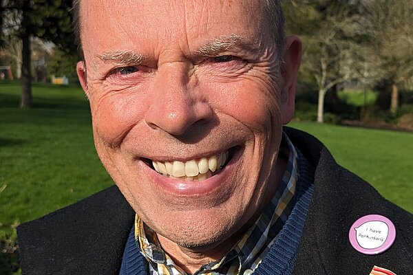 Andy Canning wearing a blue jumper, a yellow checked shirt and black suit jacket with a LGBTQ+ pride pin and a "I Have Parkinsons" pin.