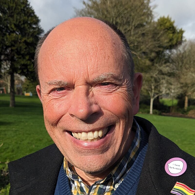 Andy Canning wearing a blue jumper, a yellow checked shirt and black suit jacket with a LGBTQ+ pride pin and a "I Have Parkinsons" pin.