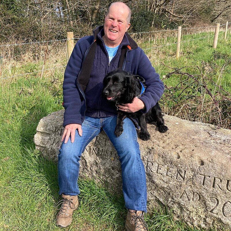 Nigel Amor sat on a log, with his black spaniel wearing a blue zipped jacket and jeans.