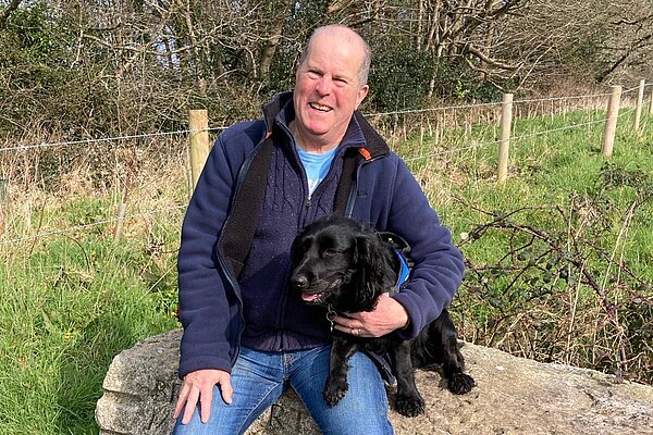 Nigel Amor sat on a log, with his black spaniel wearing a blue zipped jacket and jeans.