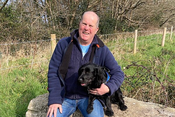 Nigel Amor sat on a log, with his black spaniel wearing a blue zipped jacket and jeans.