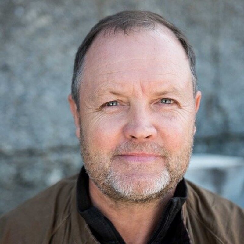 David Worthington has short hair and a grey beard. He is wearing a black top and a brown leather jacket