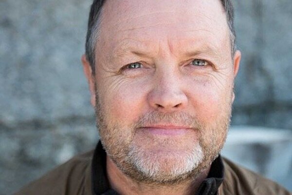 David Worthington has short hair and a grey beard. He is wearing a black top and a brown leather jacket