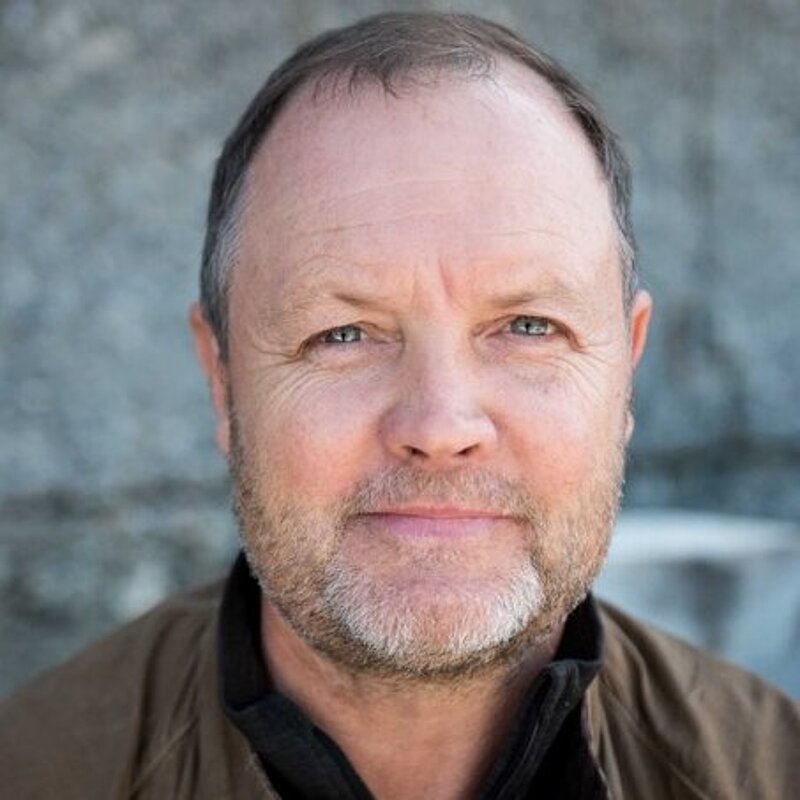 David Worthington has short hair and a grey beard. He is wearing a black top and a brown leather jacket