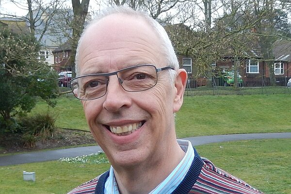 Gareth Jones is an older man with black glasses and wearing a jumper with red, white, blue, and green stripes & a light blue shirt