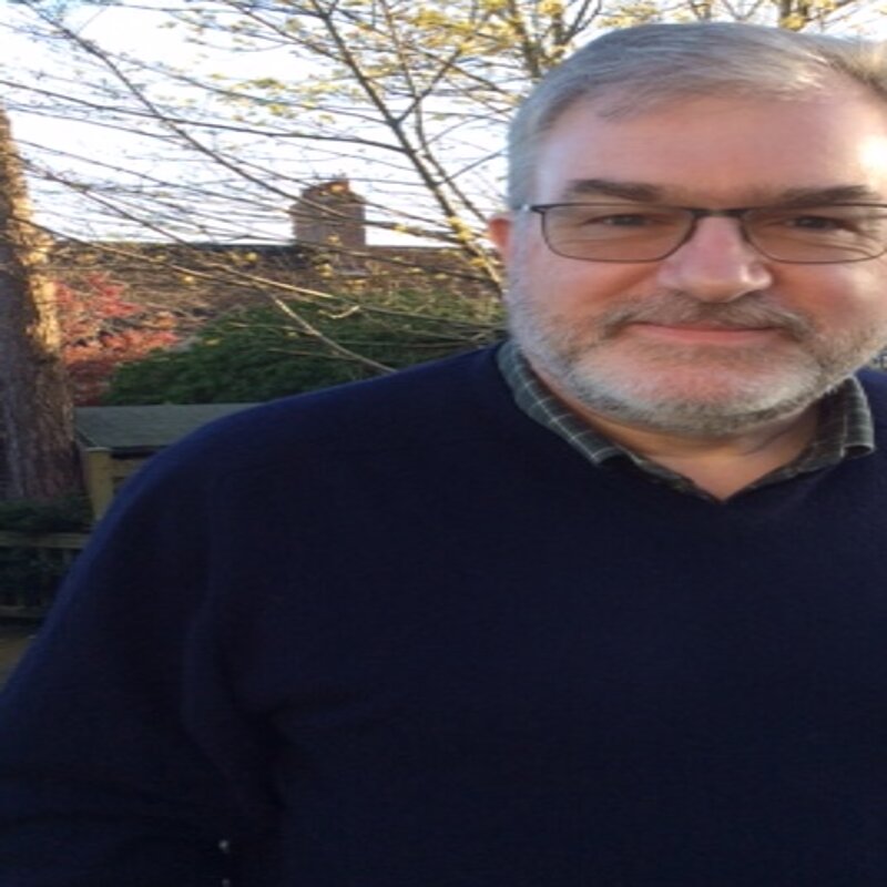 Paul Jacobs has grey hair and a beard. He is wearing a navy jumper and a green checkered shirt. He is in a garden