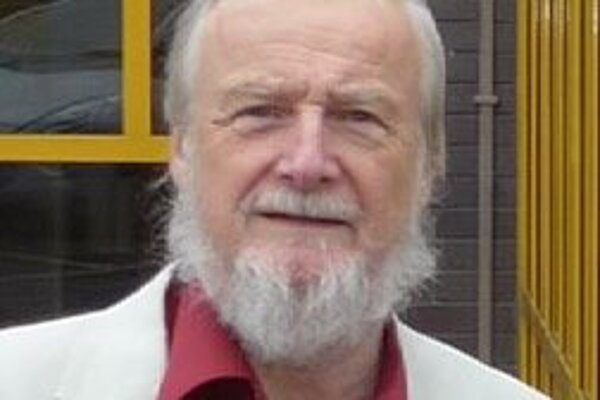 Dave Rickard is an older man with white hair and beard. In this image he is wearing a red shirt and white suit jacket
