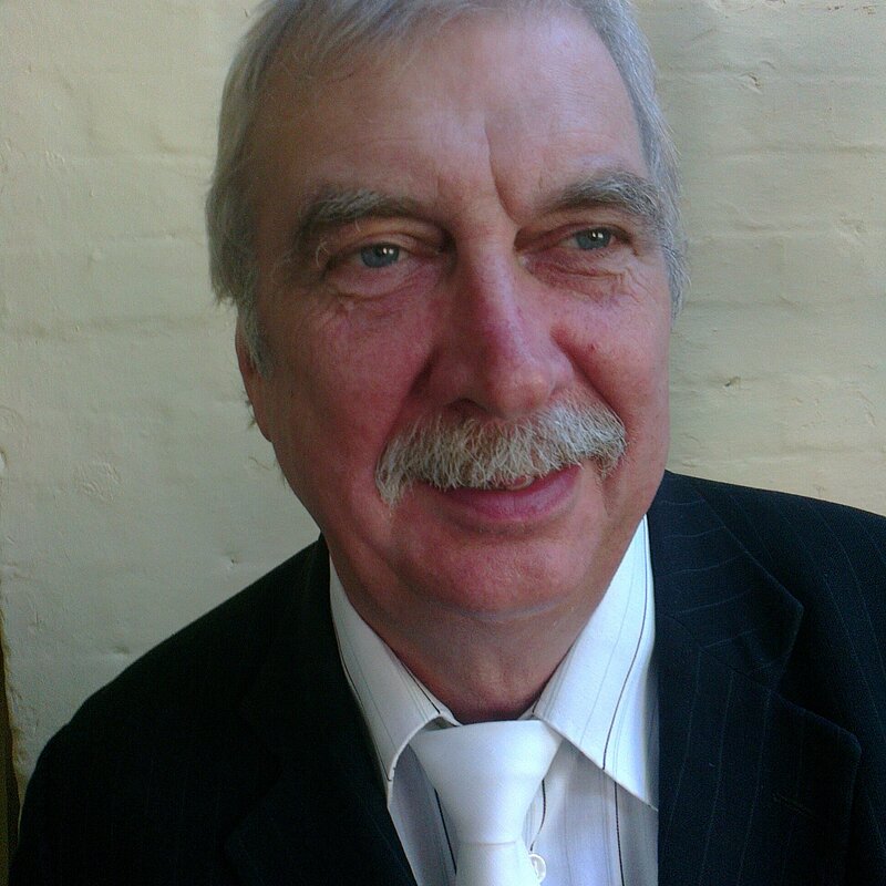 Jonathan Bourbon has grey hair and moustache. He is wearing a black suit with a white tie and white shirt
