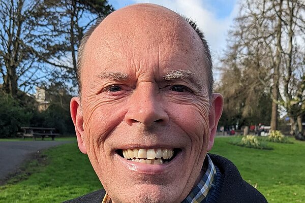 Andy Canning wearing a blue jumper, a yellow checked shirt and black suit jacket with a LGBTQ+ pride pin and a "I Have Parkinsons" pin.